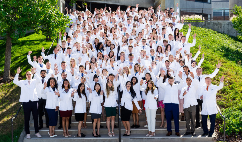 A large group of medical students in white coats gathers on outdoor steps, smiling and waving in celebration.