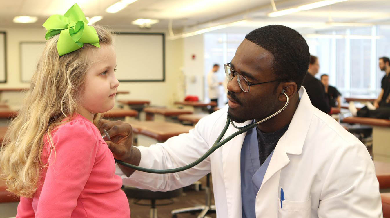 Black male student doctor uses stethoscope to check young girl's heartbeat.
