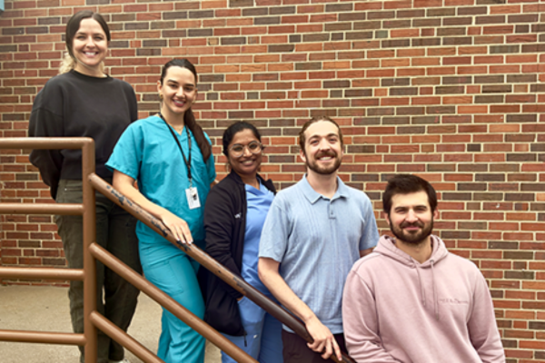 Five people stand on a staircase against a brick wall, smiling. They appear diverse and relaxed, with one in medical scrubs.