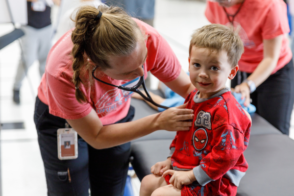 A woman in a pink shirt examines a child with a stethoscope. The child, wearing red Spider-Man pajamas, sits on a table, smiling slightly.