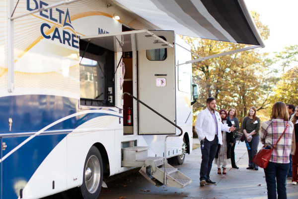 A healthcare professional speaks to a group outside a mobile clinic labeled "Delta Care-A-Van." The group is attentive and engaged. There are fall trees in the background.